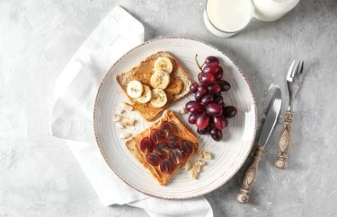 A plate with two slices of toast topped with peanut butter and slices of bananas and grapes