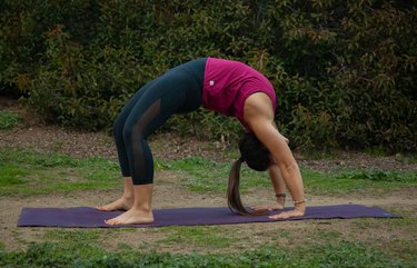 Female yoga teacher in purple top and black leggings performing backbend on a purple yoga mat in front of some trees