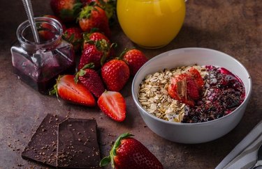 Strawberry oatmeal coated in dark chocolate in a white bowl with squares of chocolate, strawberries and a glass of orange juice