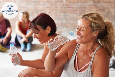 Two women laughing and drinking from water bottles after exercising to lose weight