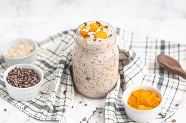 Peach Overnight Oats in mason jar on table with cloth and ingredients in bowls surrounding