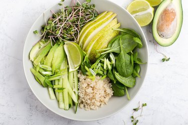 an overhead photo of a white bowl of gluten-free quinoa with spinach, sprouts, pear, lime and cucumber with an avocado and lime sliced on the counter next to the bowl