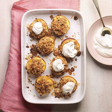 A tray of halved baked peaches, toped with almond crisp and whipped cream