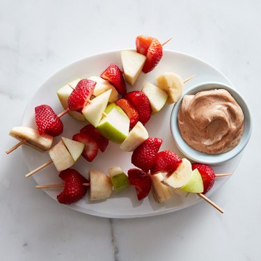 Four fruit kabobs comprised of apples, strawberries and bananas next to a peanut butter dip