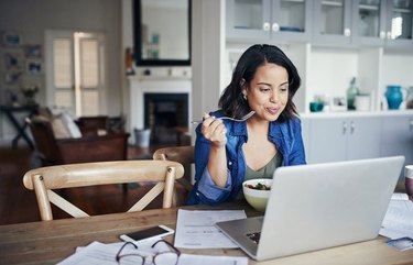 Woman eating a healthy lunch while reading on her laptop at home
