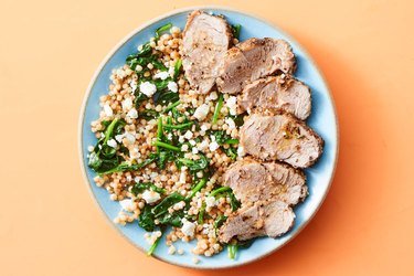 A plate of pork tenderloin with Israeli couscous, feta and spinach on a blue plate and an orange background