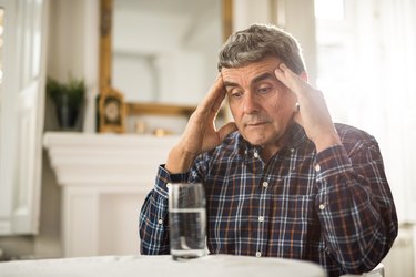 An older man sitting at a table with a glass of water, with his hands on his temples because he is feeling dizzy