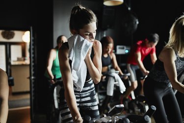 Woman wiping sweat from face with towel while riding indoor bike during cycling class in fitness studio