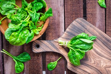 A bunch of fresh baby spinach leaves on a cutting board and spinach leaves in a bowl on a wooden table.