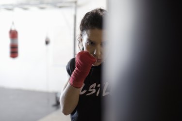 Tough, focused female boxer training at punching bag in gym
