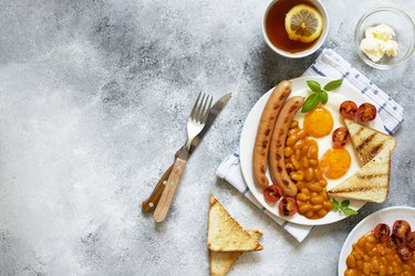 English breakfast with fried sausages, beans, mushrooms, fried eggs, grilled cherry tomatoes. Served with a cup of tea with lemon, bread toast and butter. Gray background, top view, copyspace