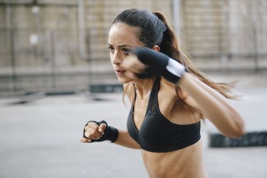 an athlete wearing a black sports bra with long hair in a ponytail and wrist wraps doing a boxing HIIT workout outside