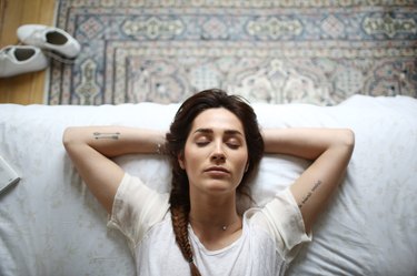 an overhead photo of a person with a long braid wearing a white t-shirt with their eyes closed and hands behind their head doing a night affirmation before going to sleep