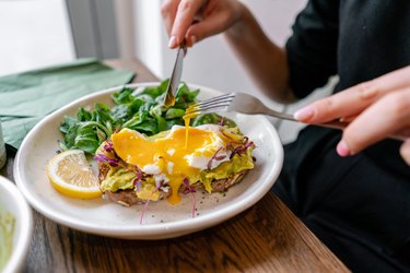 Young woman eat breakfast with knife and fork, Morning in cafe. Healthy breakfast with wholemeal bread toast with avocado, poached egg with green salad