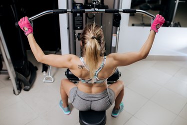Young woman working at the lat pulldown machine in the gym. Back view.