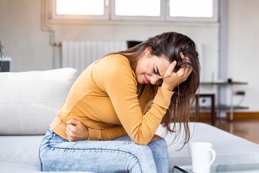 Young sick woman with hands holding pressing her crotch lower abdomen. Medical or gynecological problems, healthcare concept. Young woman suffering from abdominal pain while sitting on sofa at home