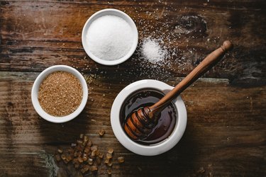an overhead photo of small bowls of white sugar, brown sugar and honey  on a dark wooden table