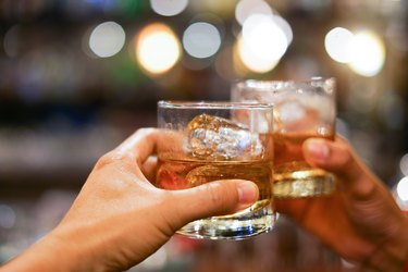 Two Hands Holding Whiskey Glasss With Ice Cubes and Whiskey In Front of Blurred Background