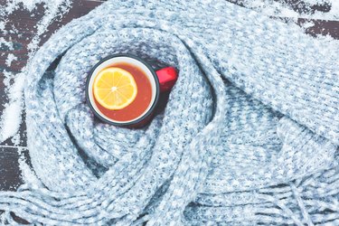 Red enameled cup of hot tea with lemon wrapped in a knitted scarf on a snowy wooden table
