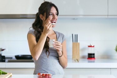 Beautiful young woman eating yogurt in the kitchen at home. Looking sideway.