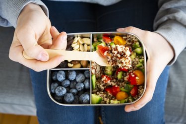 an overhead photo of a person holding a lunchbox with quinoa salad with tomato and cucumber, blueberries and trail mix