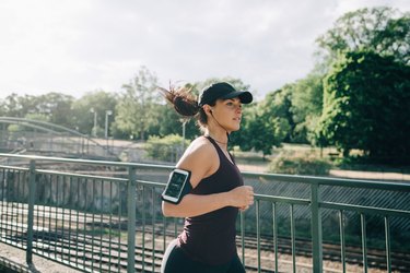 Confident sportswoman listening music through in-ear headphones while jogging on bridge in city