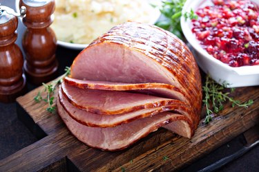 Holiday glazed sliced ham on a wooden cutting board next to cranberry sauce, mashed potatoes and salt and pepper shakers