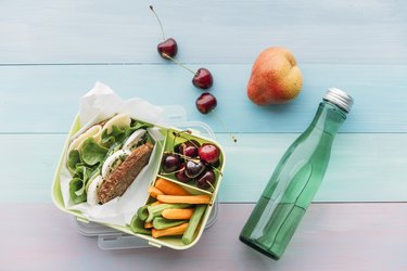 an overhead photo of a plastic container of school food next to a plastic water bottle and fruit on a blue picnic table