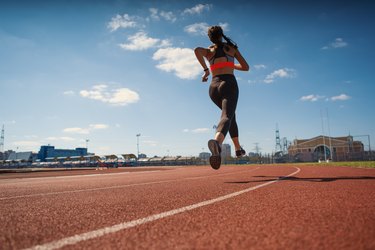 From below back view of female teen athlete in sportswear running on track at stadium on summer day