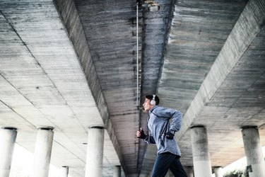 Young sporty man with headphones running under a concrete bridge in the city.