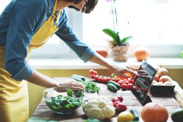 A person in a yellow apron cooking with vegetables and looking at a recipe on her tablet