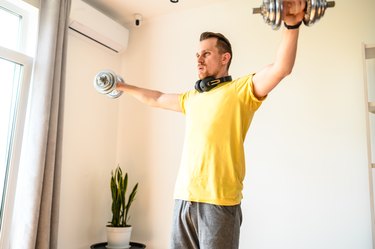 A young adult in a yellow t-shirt and gray shorts does a lateral dumbbell raise at home for stronger shoulders