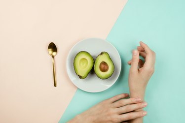 Flatlay with cut avocado on white plate and woman's hands on pastel background, healthy diet concept