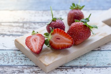 Two half cut and whole strawberries with leaves on cutting board on vintage table