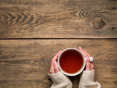 Female hands holding a mug of hot black tea. Cold winter white warm clothes. Top view. Aged rustic wooden background