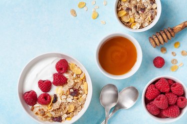 Greek yogurt in bowl with ingredients raspberries, honey and muesli on blue table top view.