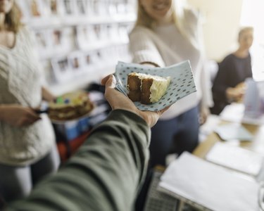 a coworker wearing a green long sleeve shirt hands a slice of cake at an office birthday celebration to a person with long blonde hair wearing a white sweater