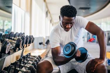 Young man sitting and lifting a dumbbell with the rack at gym