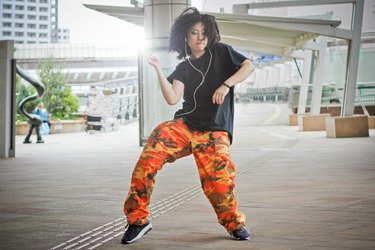a young adult wearing orange camo pants and a black t-shirt dances outside listening to music on headphones for stress relief
