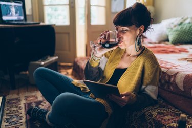 A happy woman drinking a glass of wine at home while using her tablet