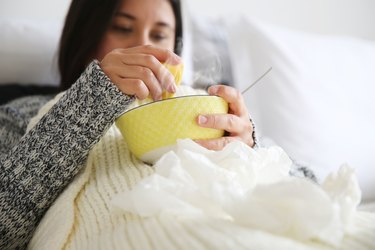 Sick person wearing a grey sweater and a white blanket drinking soup for a sore throat