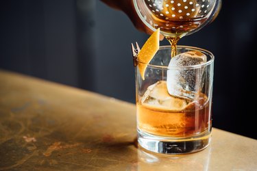 closeup of a cocktail shaker pouring into glass with dark alcohol, ice and orange peel