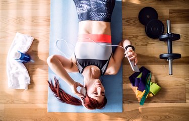 Woman lying on exercise mat listening to music on her smartphone while working out at home