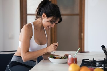 Sporty young woman eating salad and drinking fruit juice in the kitchen at home.