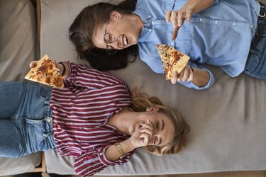 Two laughing young women lying down eating pizza together