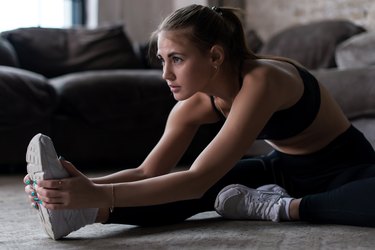 Slim sporty girl sitting on floor doing stretching exercise indoors