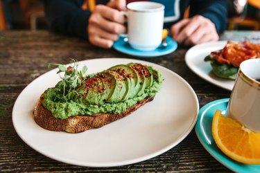 Sandwich or toast with avocado in the foreground. A person is going to eat it and drink coffee or tea
