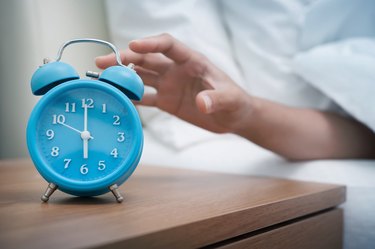 A person's hand hitting the snooze button on a blue alarm clock next to the bed