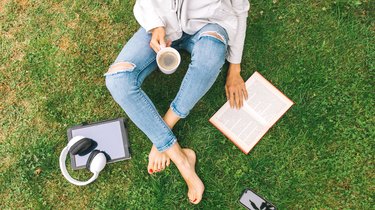 Young woman sitting on the grass drinking coffee and reading a book