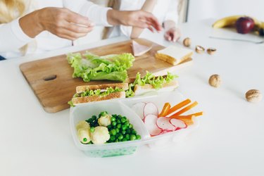 Snack meal prep with sandwich and fresh vegetables, bottle of water on the home kitchen table. In the background, the mother and her daughter are preparing food. Healthy eating concept